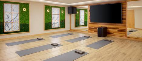 Una habitación con suelo de madera, ventanas amplias con follaje verde, un televisor de pantalla plana y colchonetas de yoga dispuestas en el suelo.