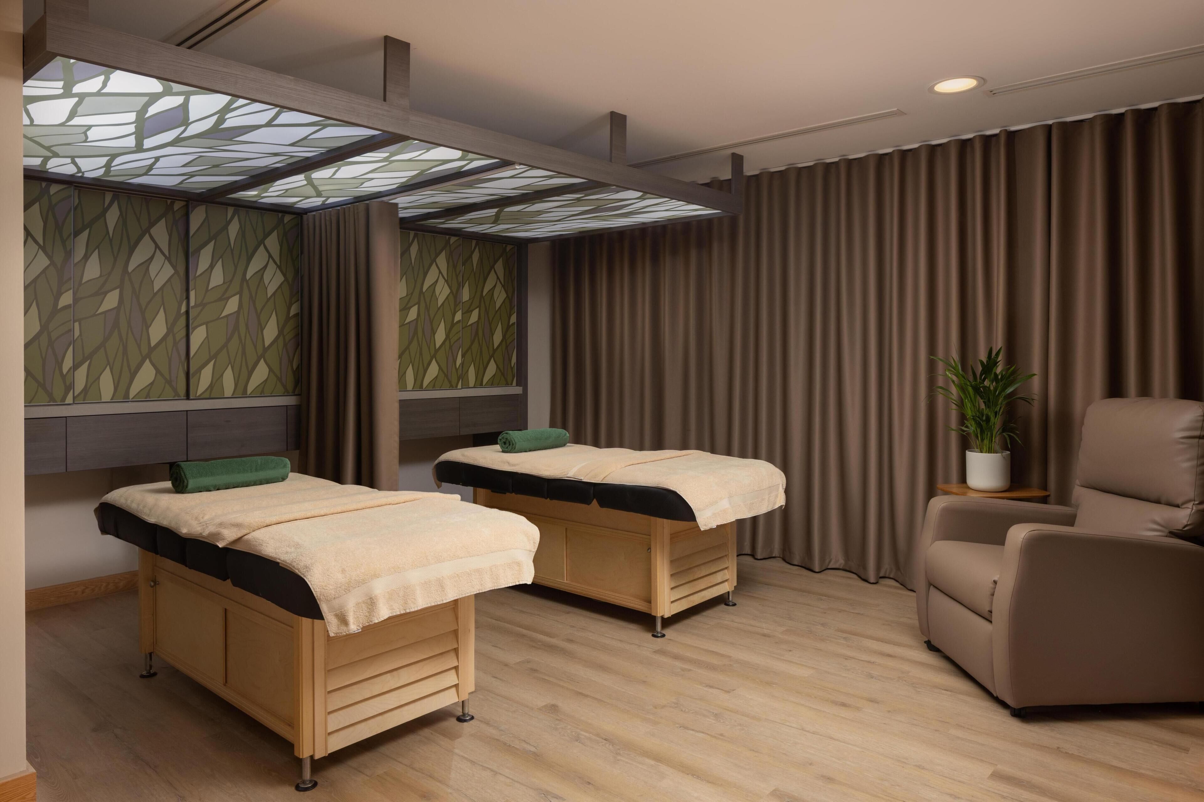 Ett rum med två sängar, en massagefåtölj, en krukväxt och ett stort fönster med ett geometriskt mönster.
