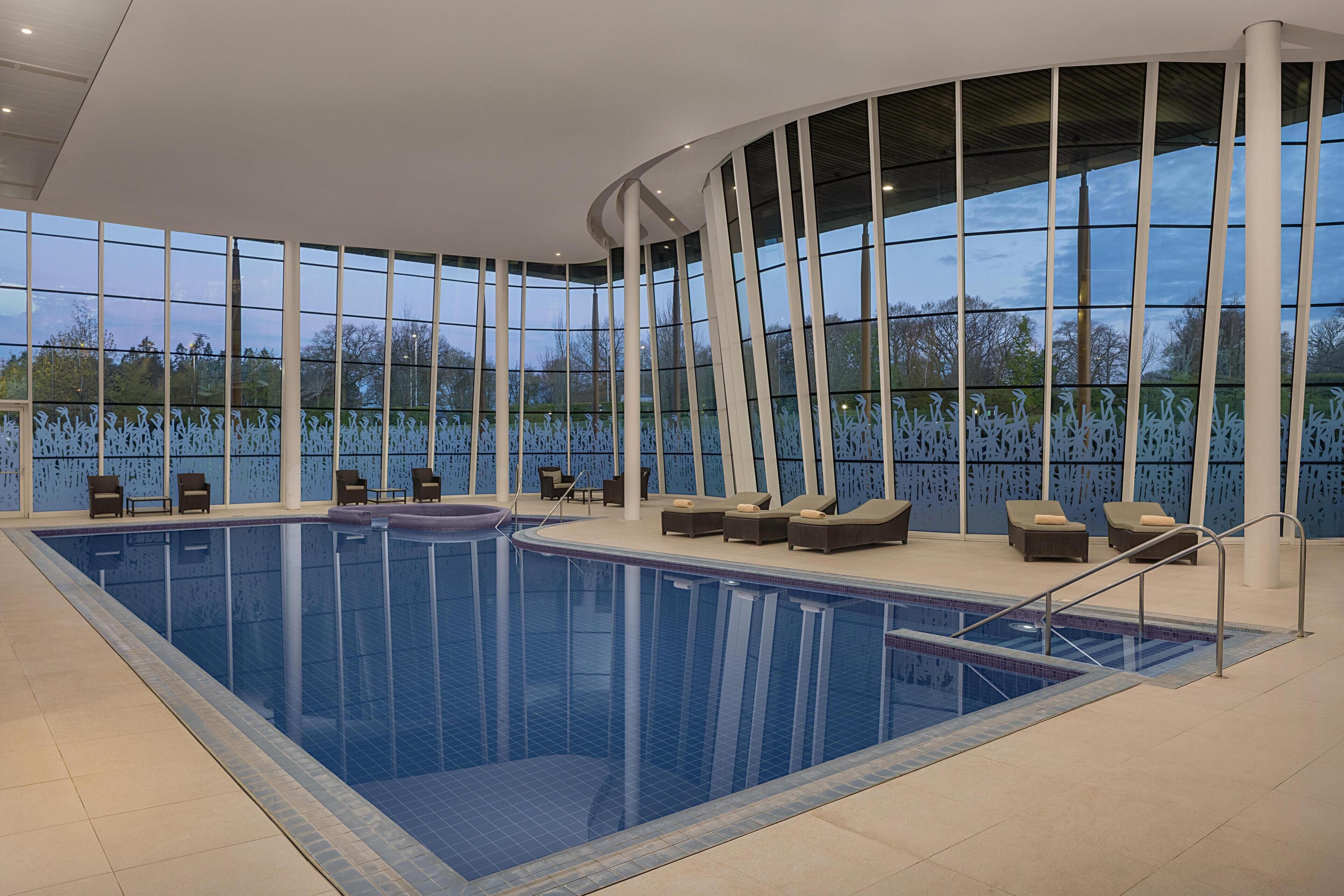 Piscina interior moderna com paredes de vidro transparente, cadeiras de lounge e vista para o exterior.