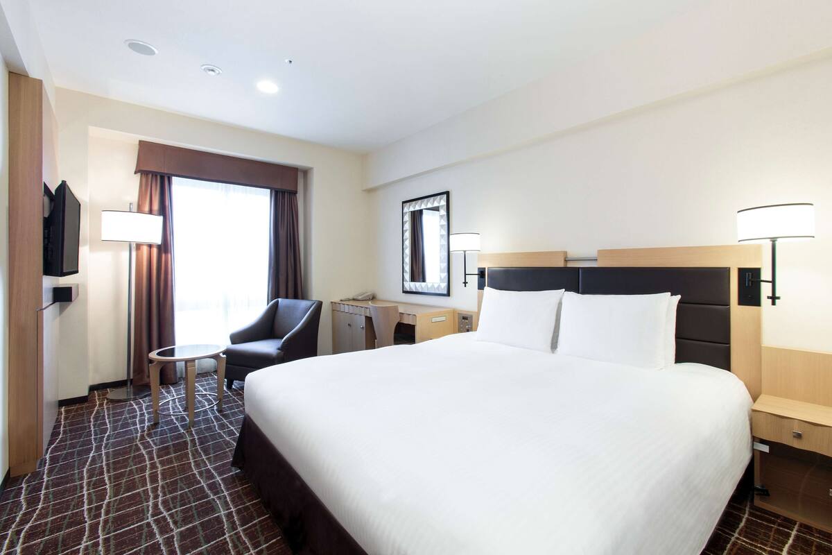 Una habitación de hotel con una cama grande, un escritorio, una silla, un televisor y una ventana con cortinas.