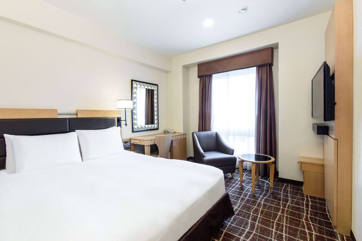 Una habitación de hotel con una cama grande, un escritorio, una silla, una mesita, un televisor y una ventana con cortinas.