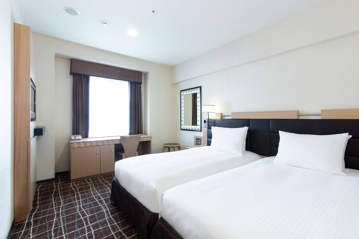 Habitación de hotel con dos camas, un escritorio, una silla, un ventanal con cortinas, un espejo y una lámpara.