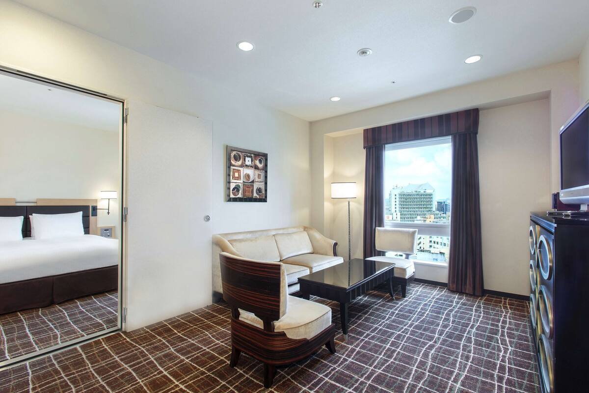 Habitación de hotel con cama, sofá, sillón, mesita, lámpara, ventana con cortinas y lavadora.