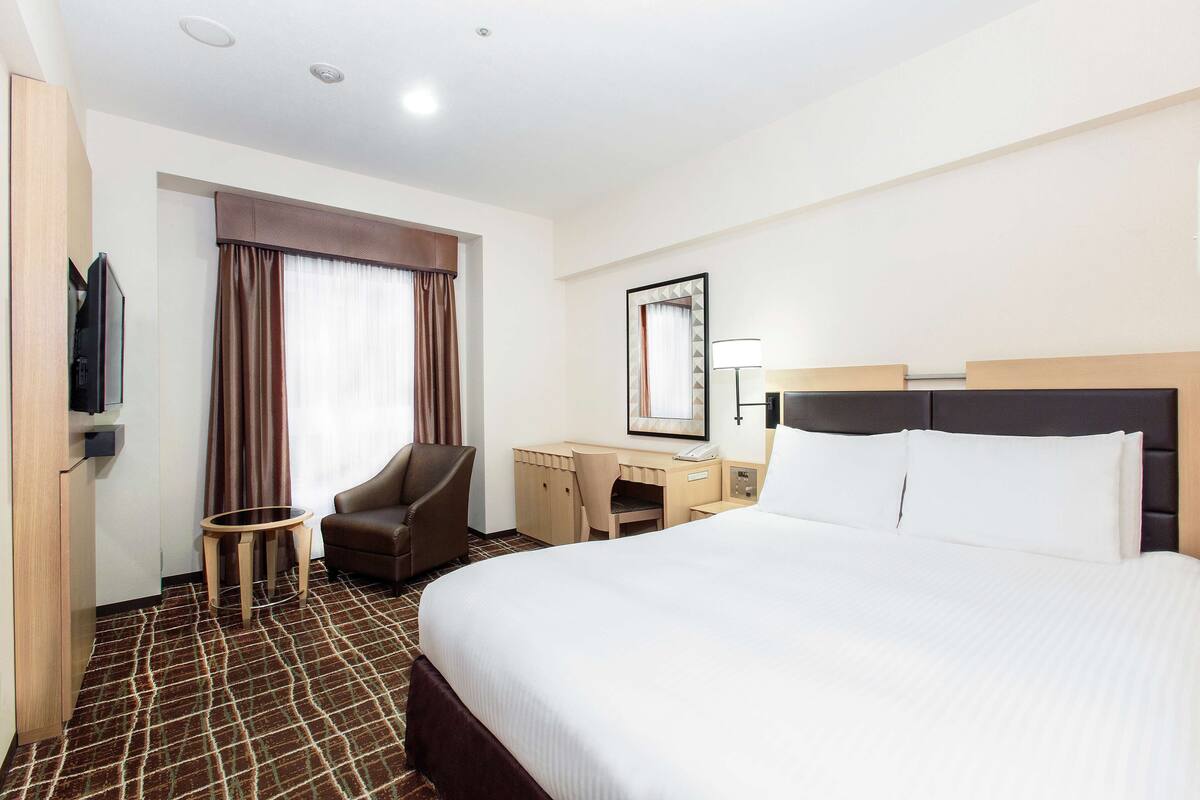 Habitación de hotel con una cama grande, un escritorio con silla, un televisor y una ventana con cortinas.