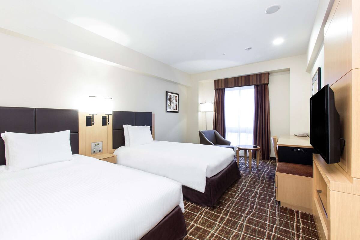 Habitación de hotel con dos camas, un escritorio, una silla, un televisor y una ventana con cortinas.