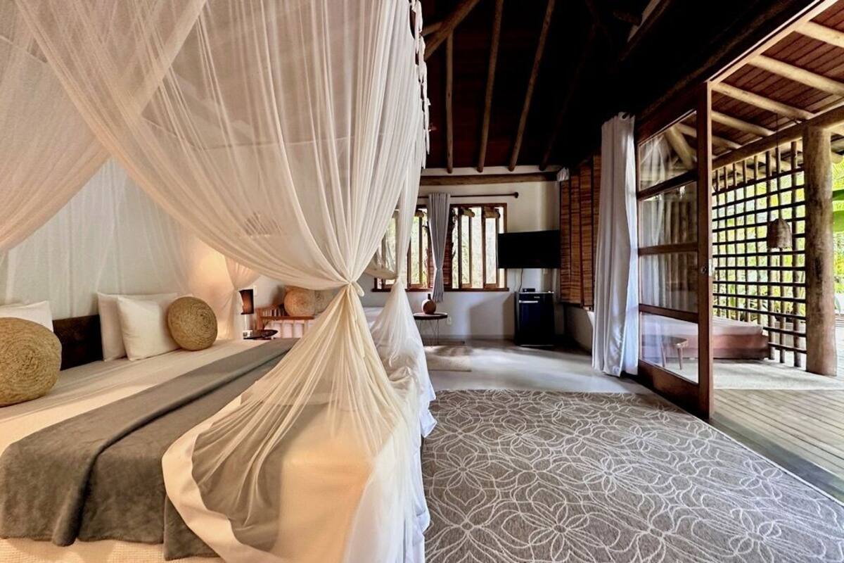 A room with a bed under a canopy, a patterned rug, a wooden ceiling, and a view through a lattice window.