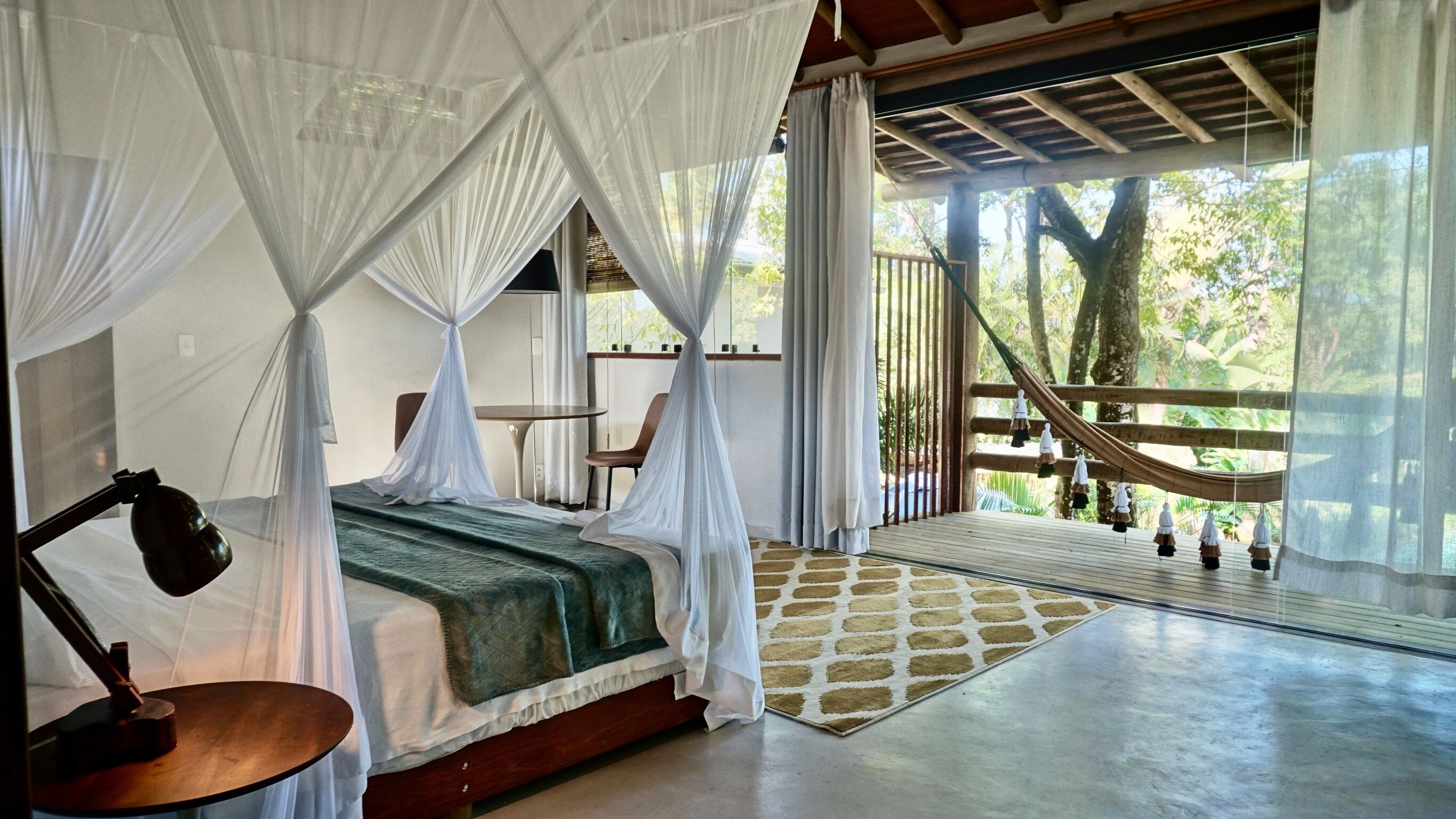 A bedroom with a bed covered by a white canopy, a bedside table with a lamp, and a view of a hammock outside.