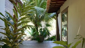 A wooden deck with potted plants and a palm tree, surrounded by a building with large windows. - Soleluna Casa Pousada (Porto Seguro)
