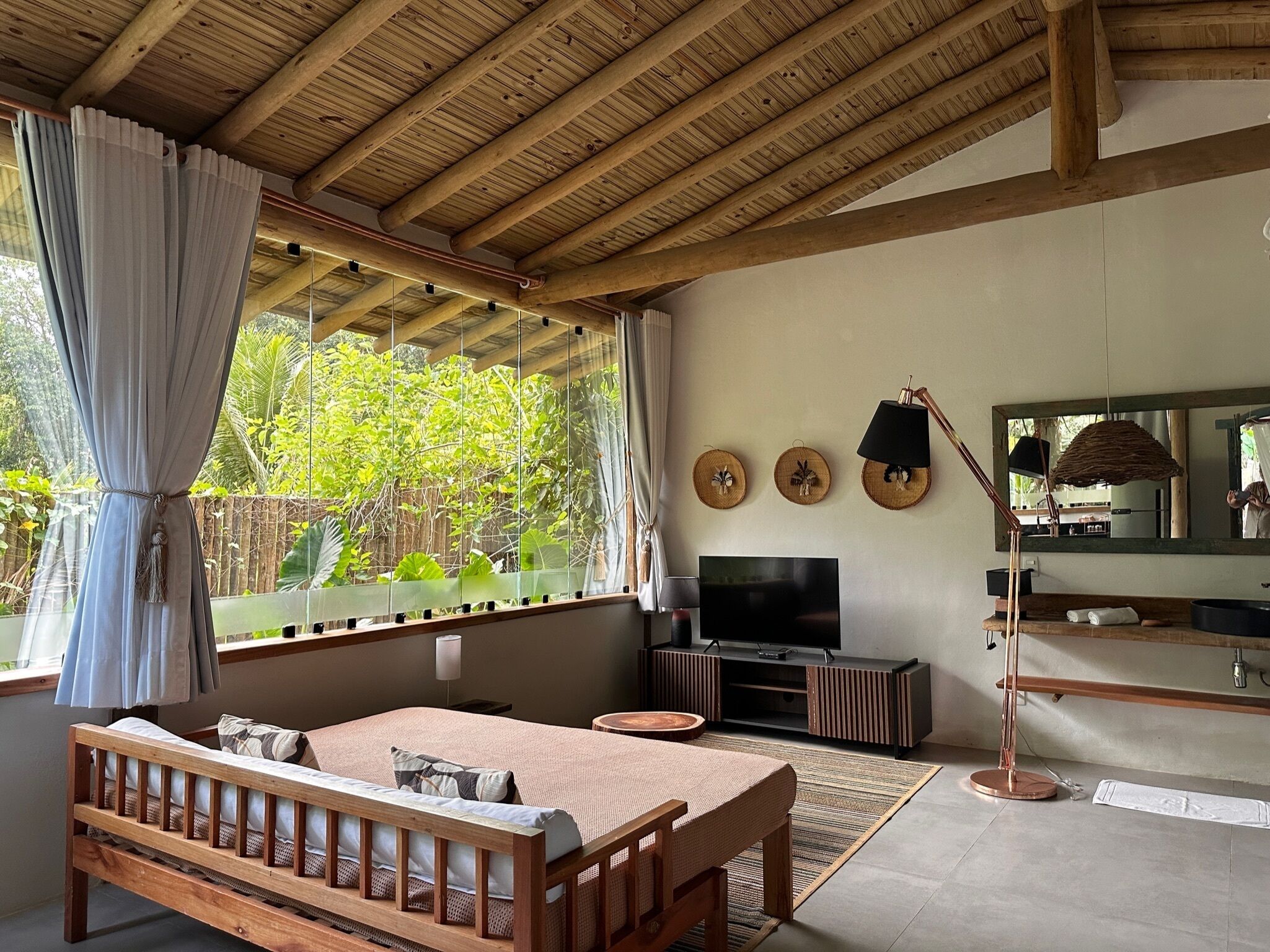 A bedroom with a wooden ceiling, a bed, a TV, a mirror, and a window with curtains.