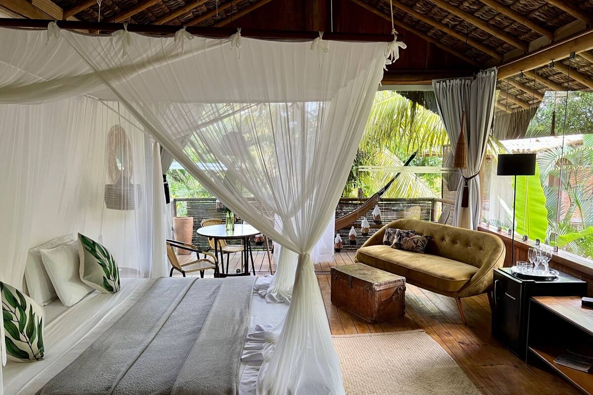 A room with a bed, a sofa, a table, and a hanging chair. There is a window with curtains and a view of greenery outside.