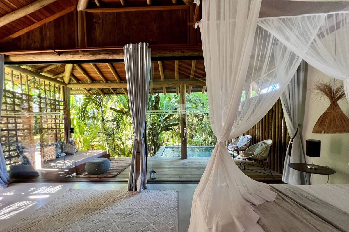 A room with a bed, a sitting area with cushions, a small table, and a view of a pool and greenery.
