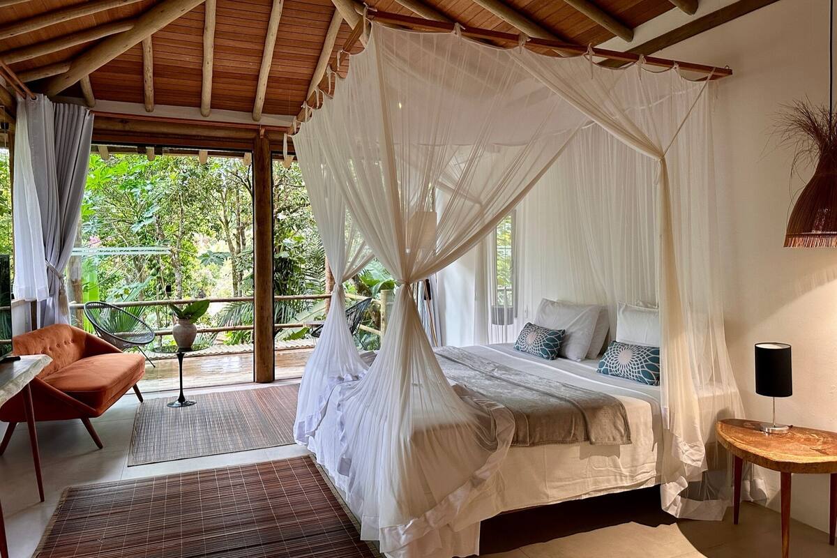 A bedroom with a canopy bed, a chair, a small table, and a view of greenery outside.