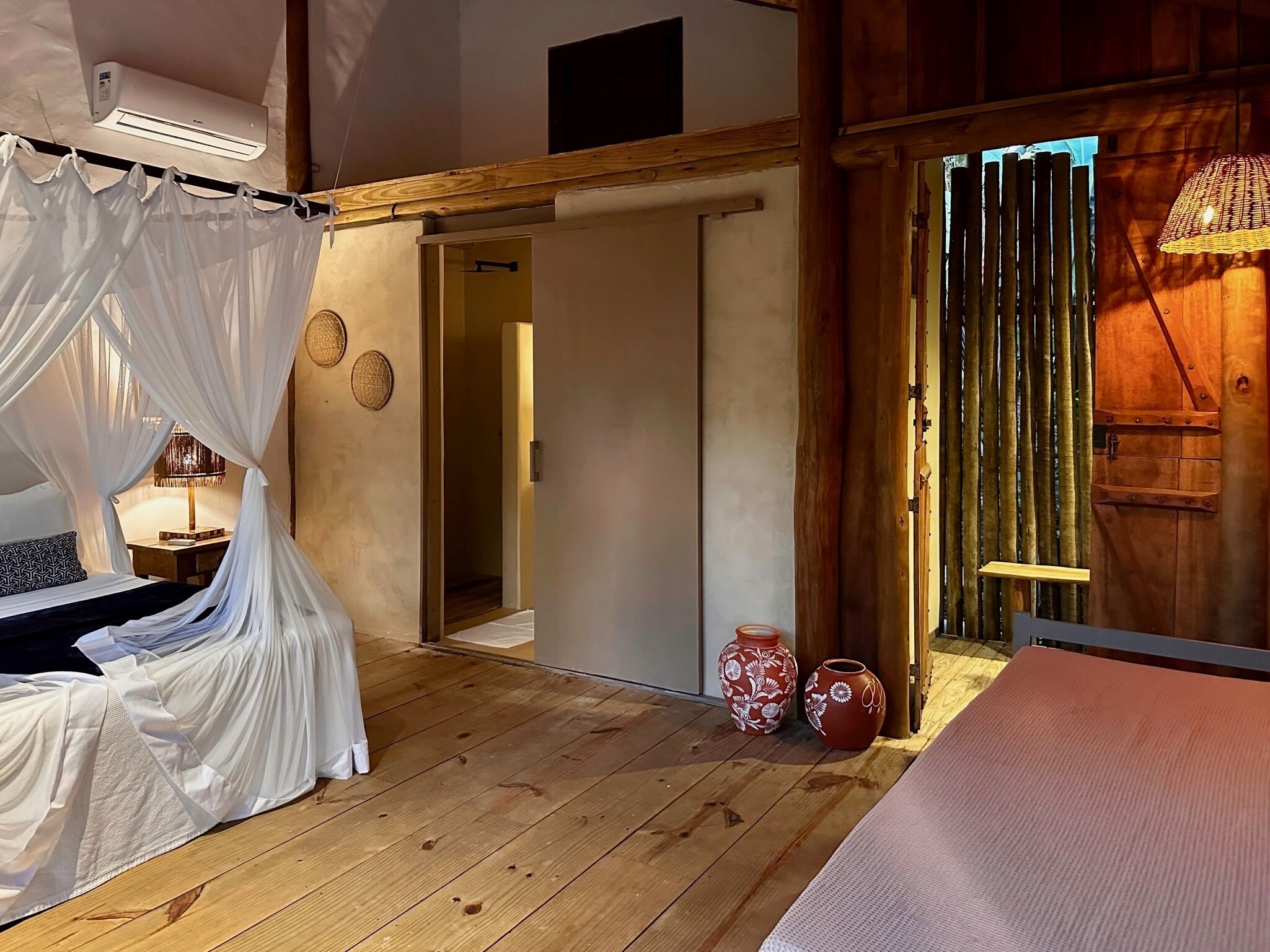 A bedroom with a wooden floor, a bed with white curtains, a bedside lamp, and two decorative vases.