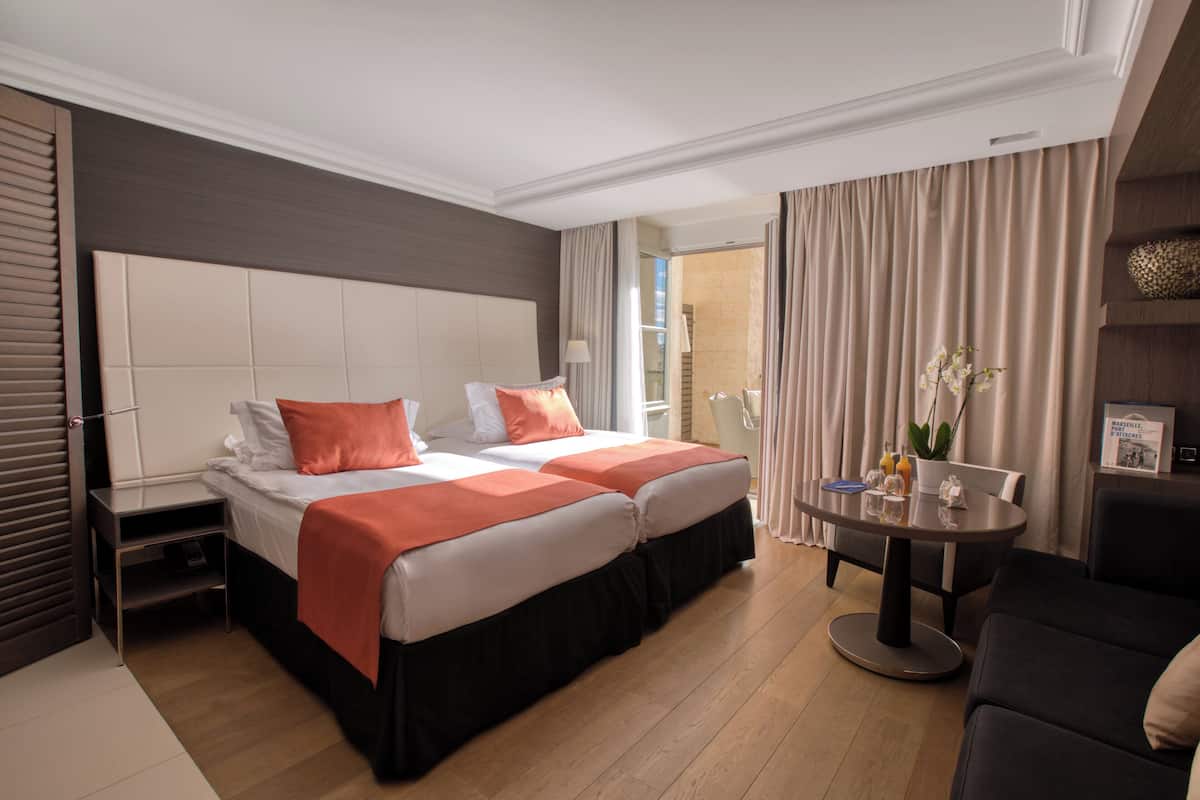 Une chambre d’hôtel avec deux lits, une table ronde, un canapé et une grande fenêtre avec des rideaux.
