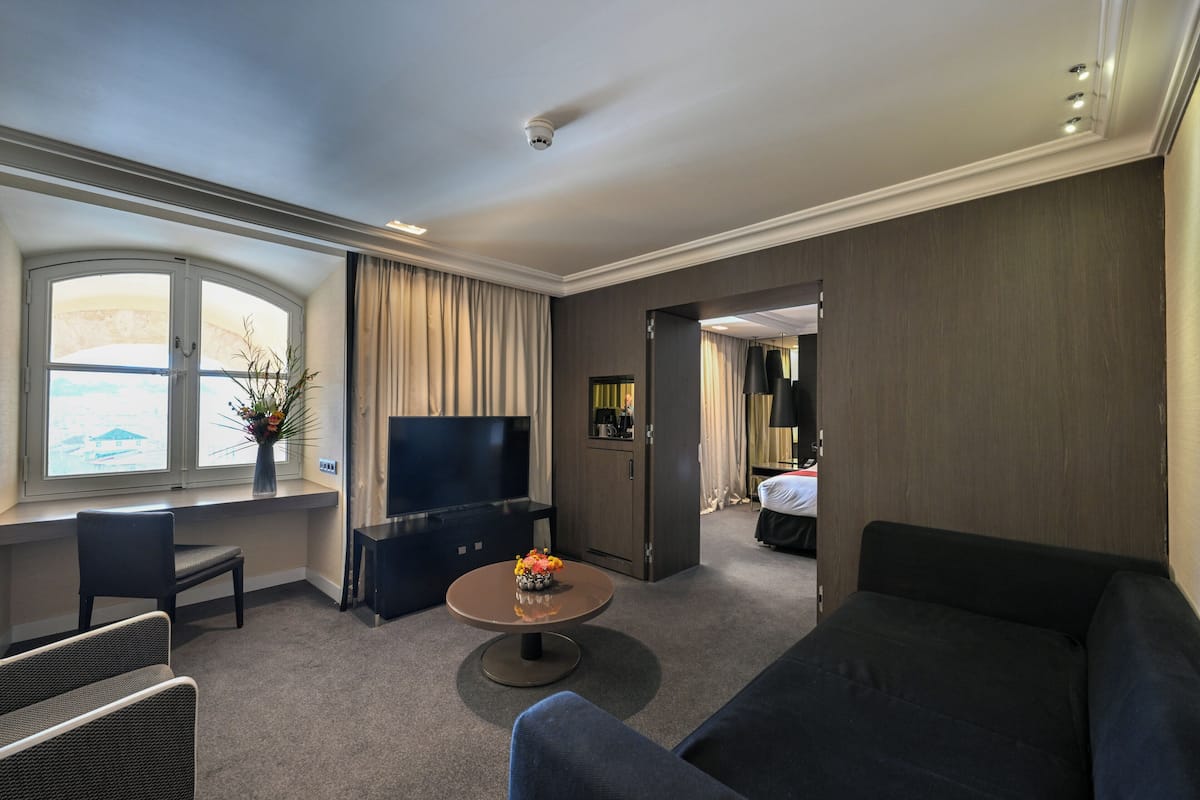 Une chambre d’hôtel avec un canapé, une télévision, une petite table avec un vase, une fenêtre avec des rideaux et une porte donnant sur une autre pièce.