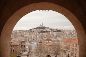 Vista de un paisaje urbano a través de un ventanal arqueado, con una colina destacada que tiene un edificio en la cima, rodeada de edificios muy juntos y una masa de agua al frente.