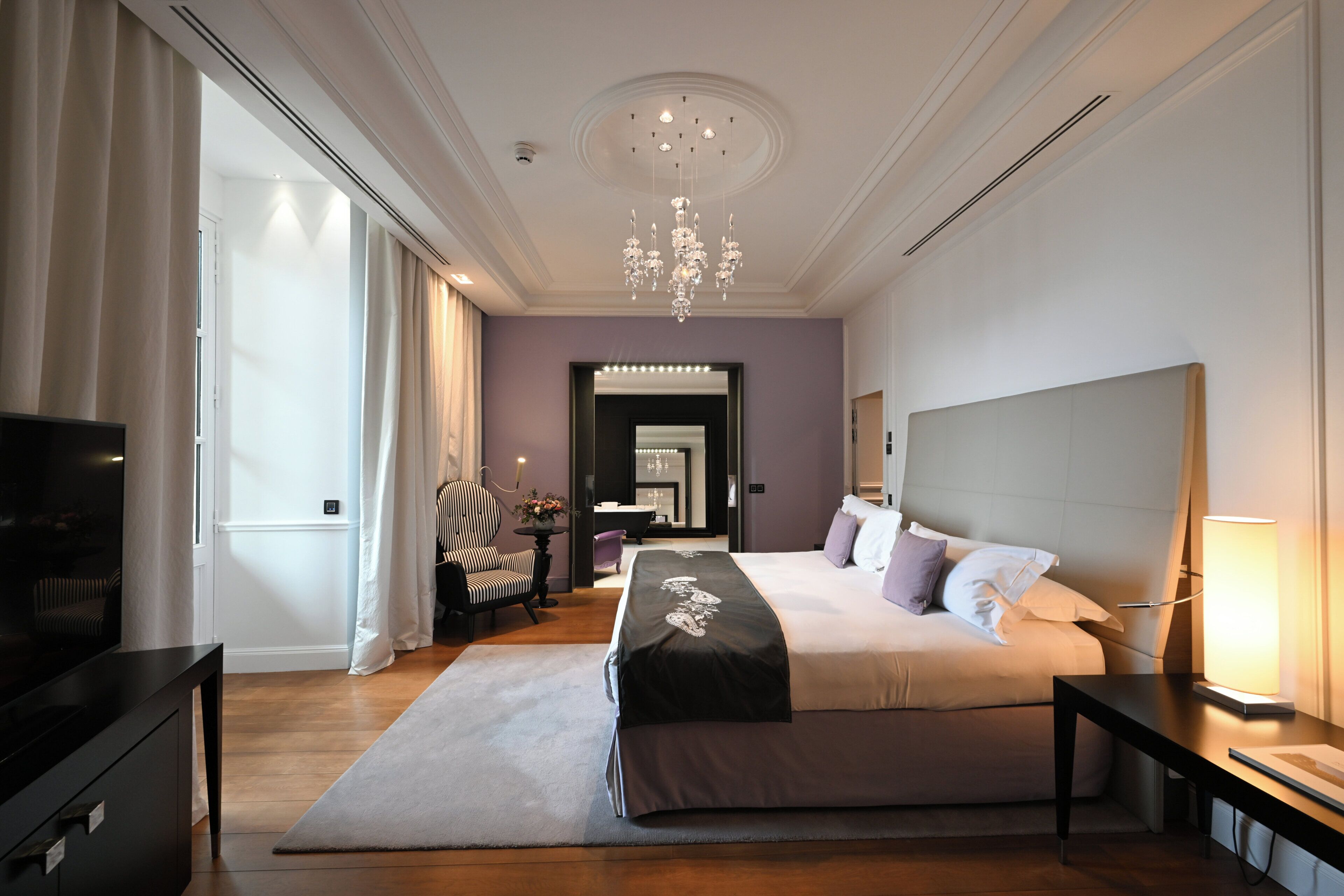 A modern hotel room with a large bed, a television, a desk, and a chandelier.