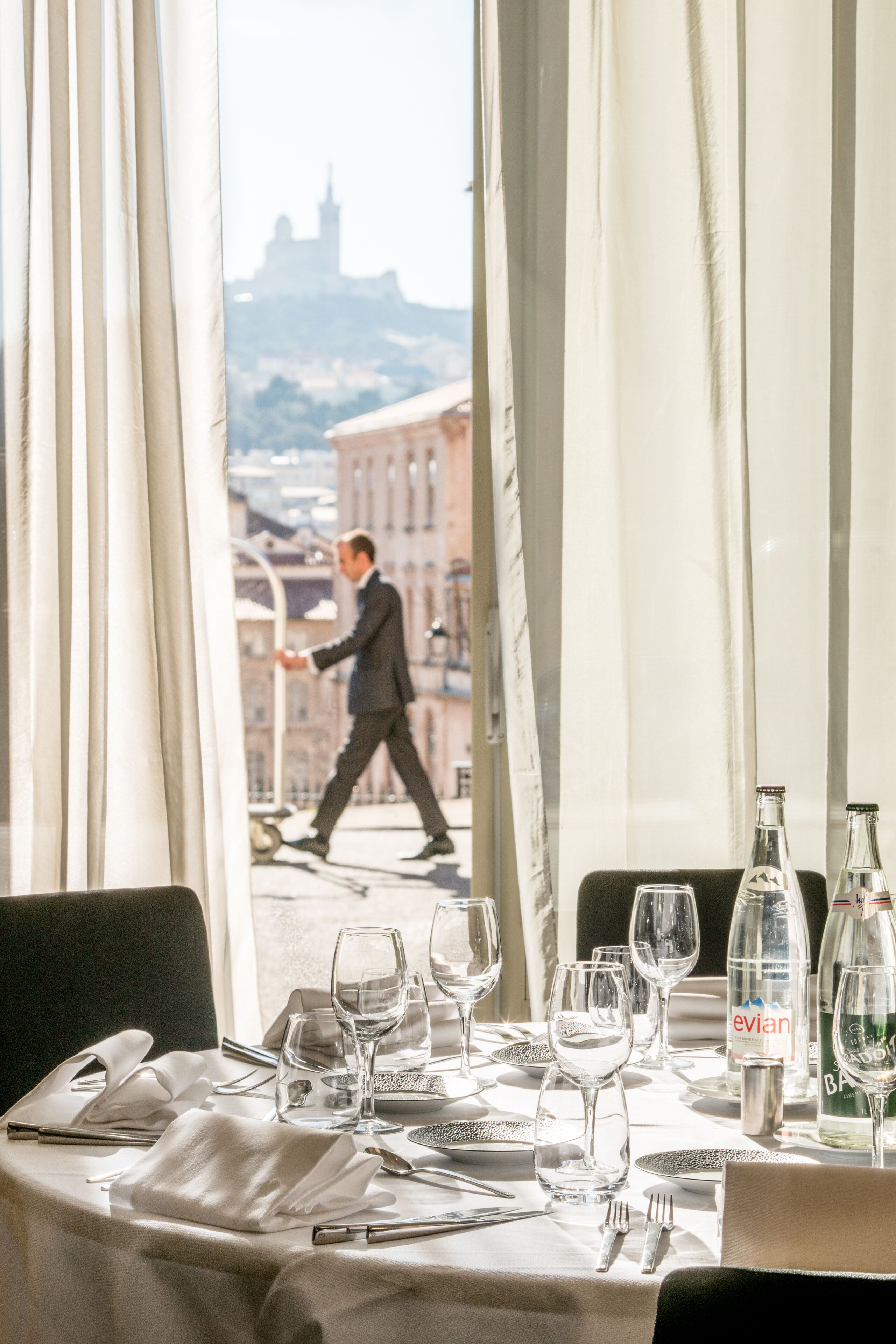Une table dressée pour le dîner, avec des bouteilles de vin et des verres, une vue sur la ville à travers une fenêtre, et une personne qui marche à l’extérieur.