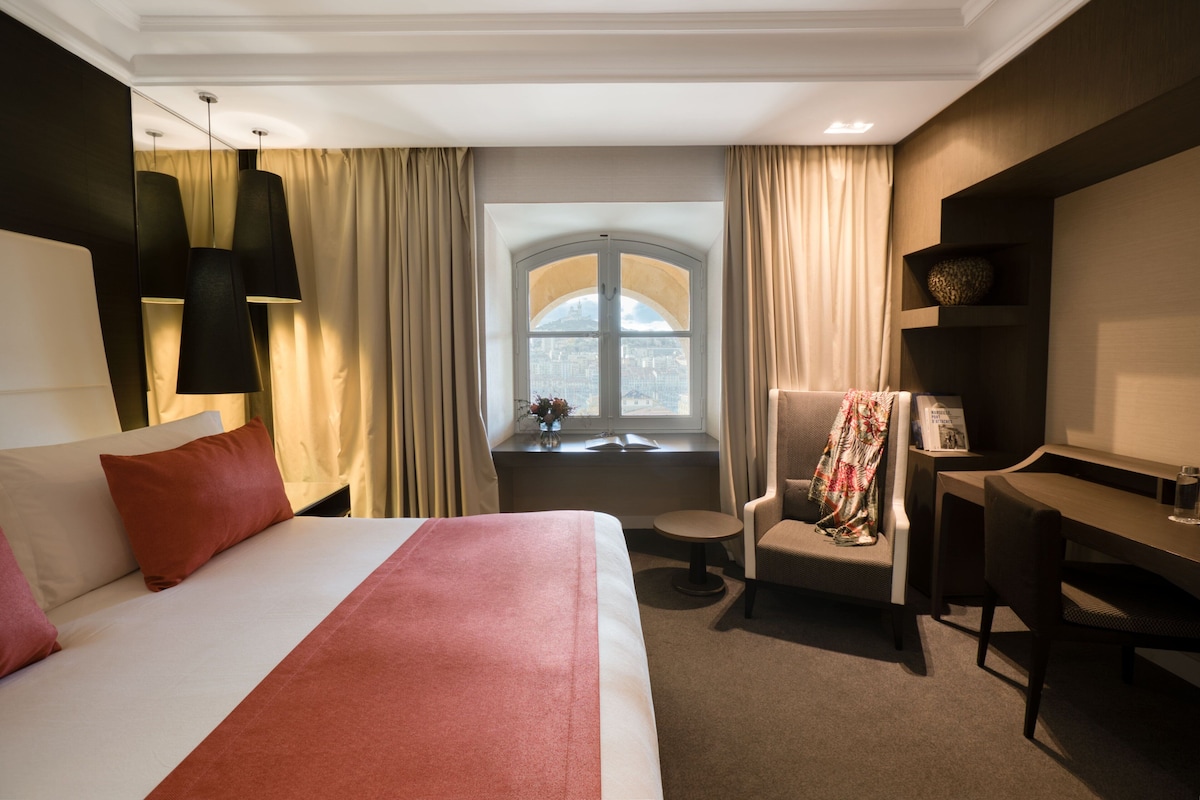 Una habitación de hotel con una cama grande, un escritorio, una silla y un ventanal con cortinas.