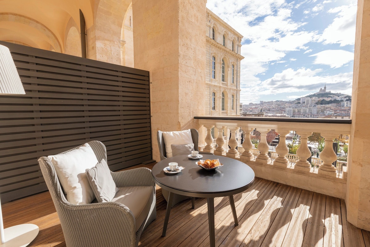 Un balcón con mobiliario de mimbre, una mesa con un cuenco y un platillo, y vistas de un paisaje urbano con edificios históricos.