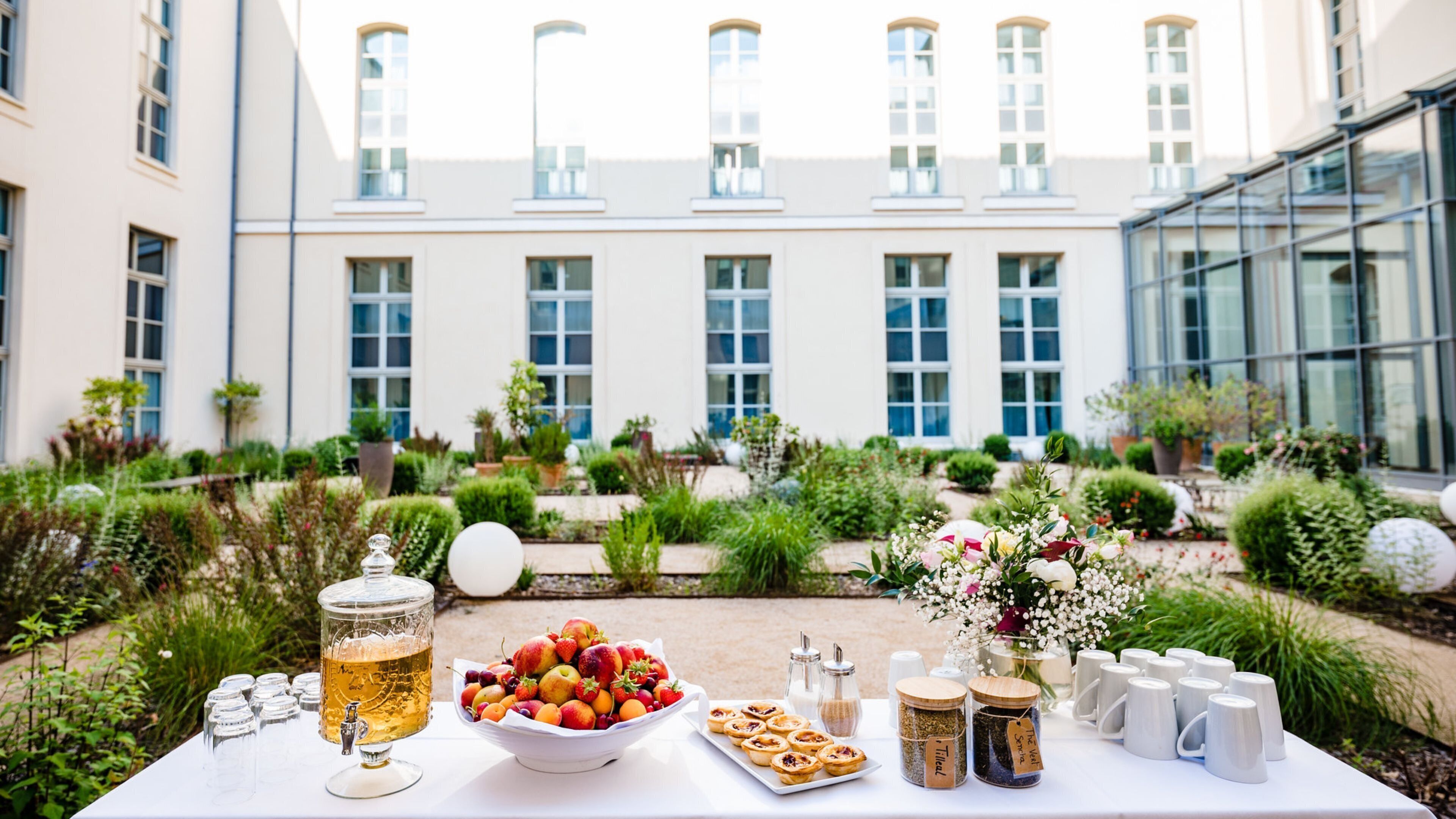 Uma mesa com um dispensador de bebidas, uma cesta de frutas, doces e potes de chá, montada ao ar livre com um edifício e um jardim ao fundo.