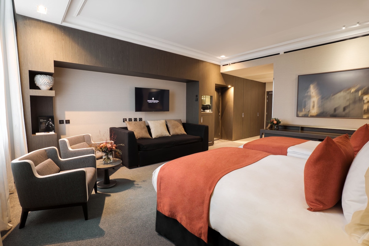 Una habitación de hotel moderna con una cama grande, dos sillones, un sofá, un televisor de pantalla plana y un cuadro en la pared.