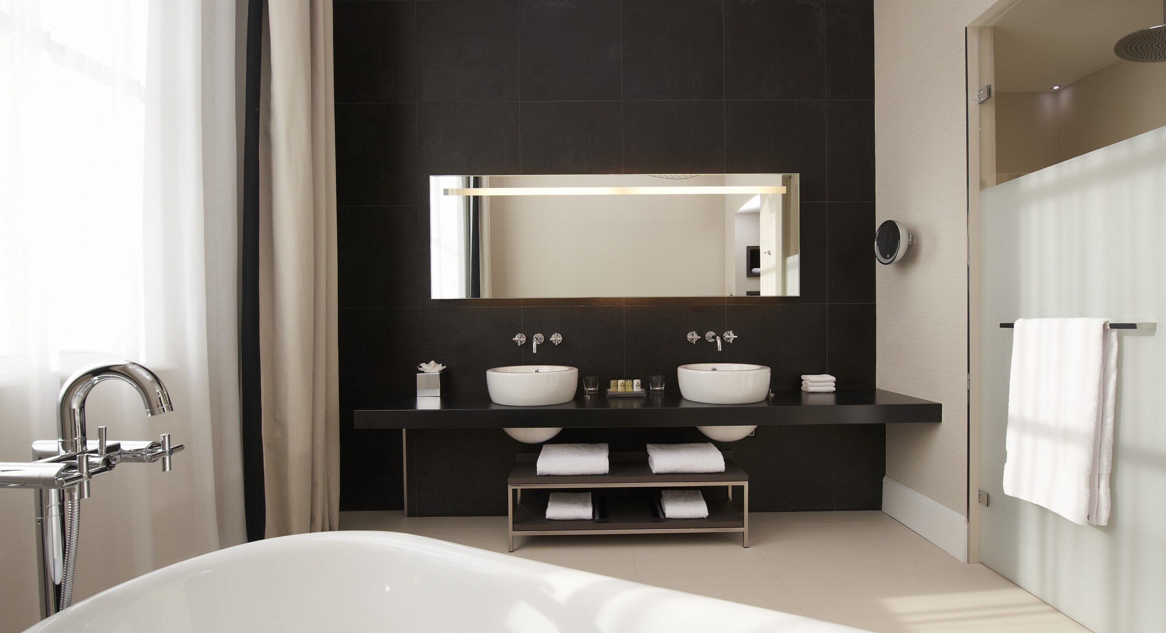 A modern bathroom with a double sink, a large mirror, a bathtub, and a towel rack.