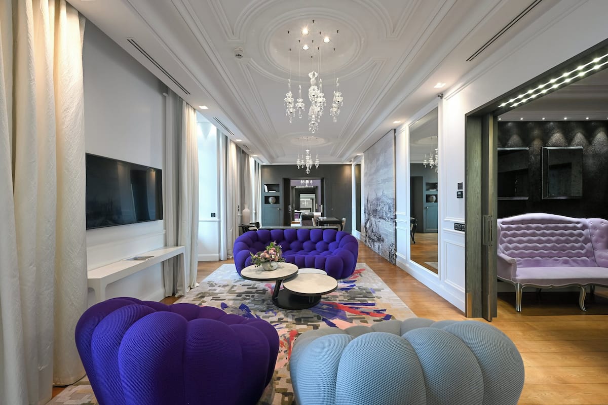 Un pasillo amplio con una zona de estar grande que incluye sofás morados y azules, un candelabro y un televisor de pantalla plana montado en la pared.