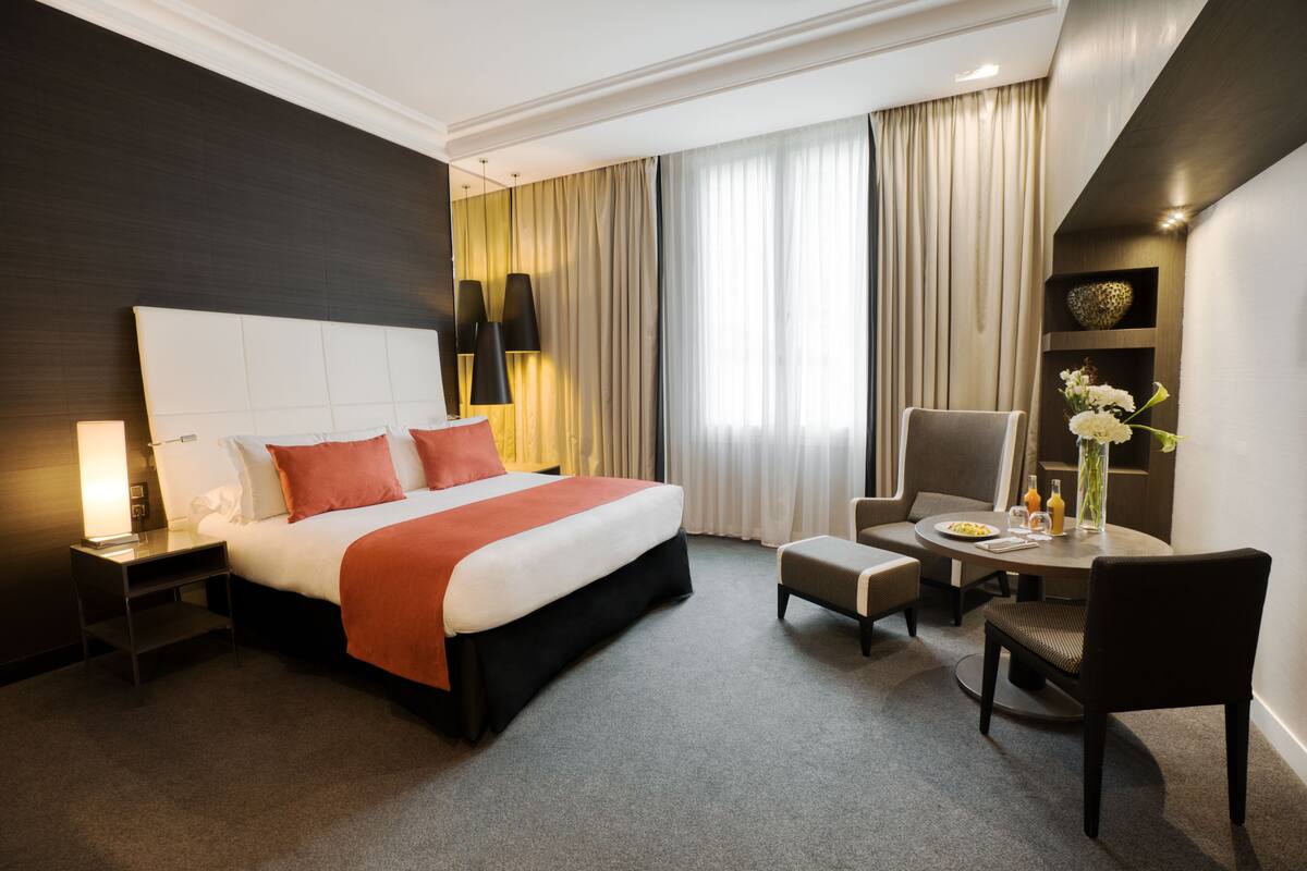 Une chambre d’hôtel avec un grand lit, un bureau avec une chaise, une petite table avec un vase et des rideaux.