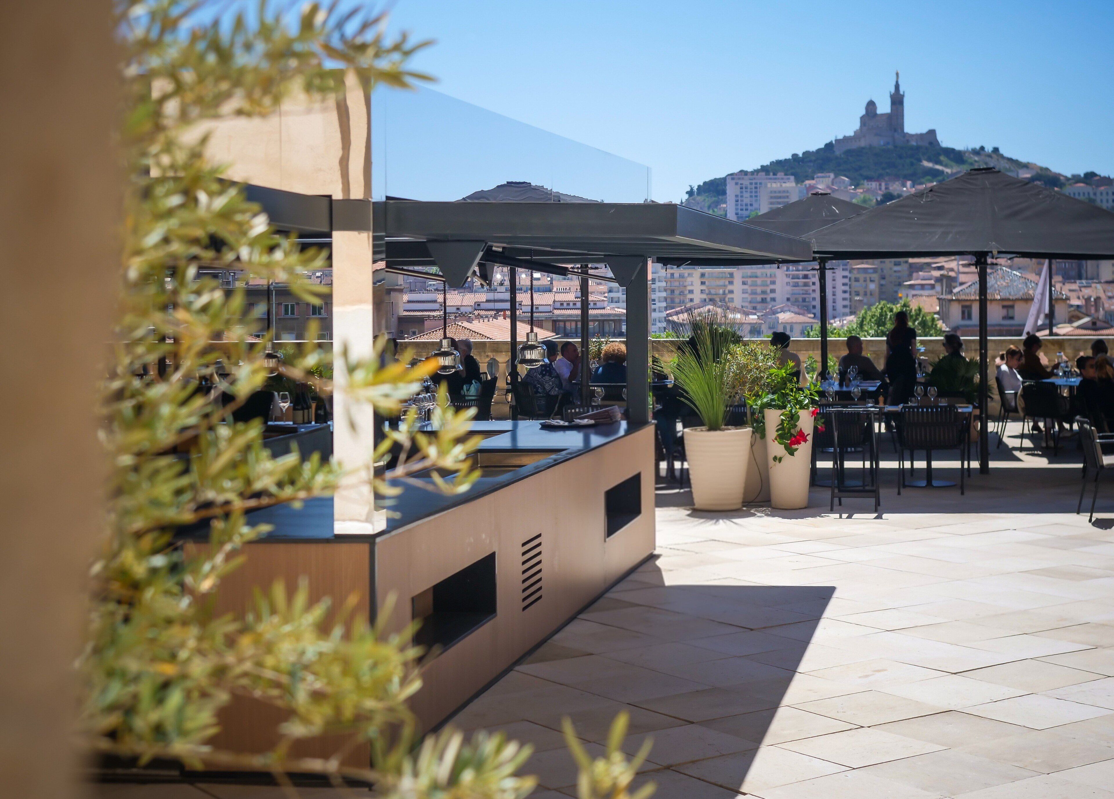 An outdoor dining area with tables and chairs, potted plants, and a view of a hill with buildings.