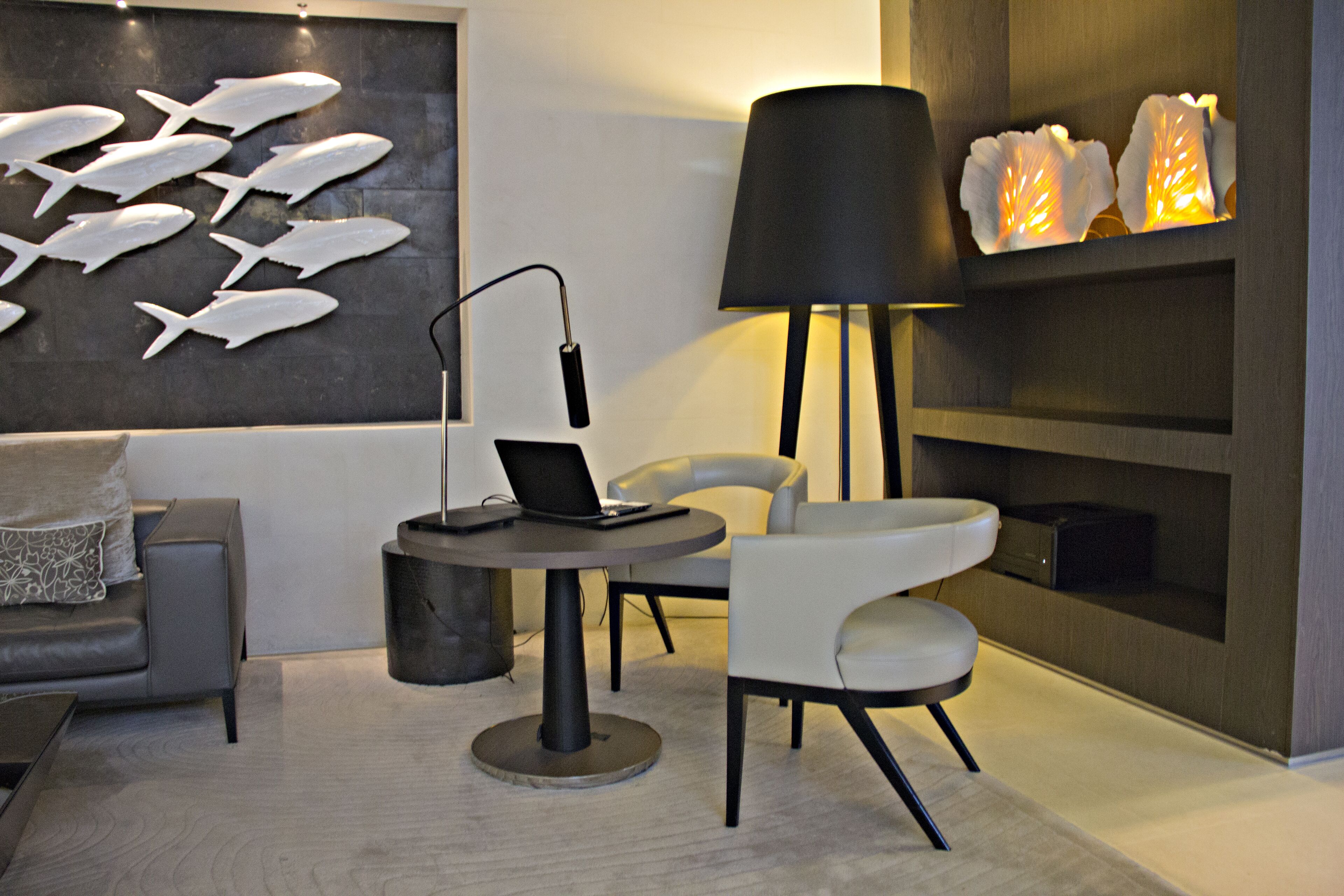 A modern living room with a grey sofa, a round table with a laptop, a black floor lamp, and a wall with fish-shaped decorations.