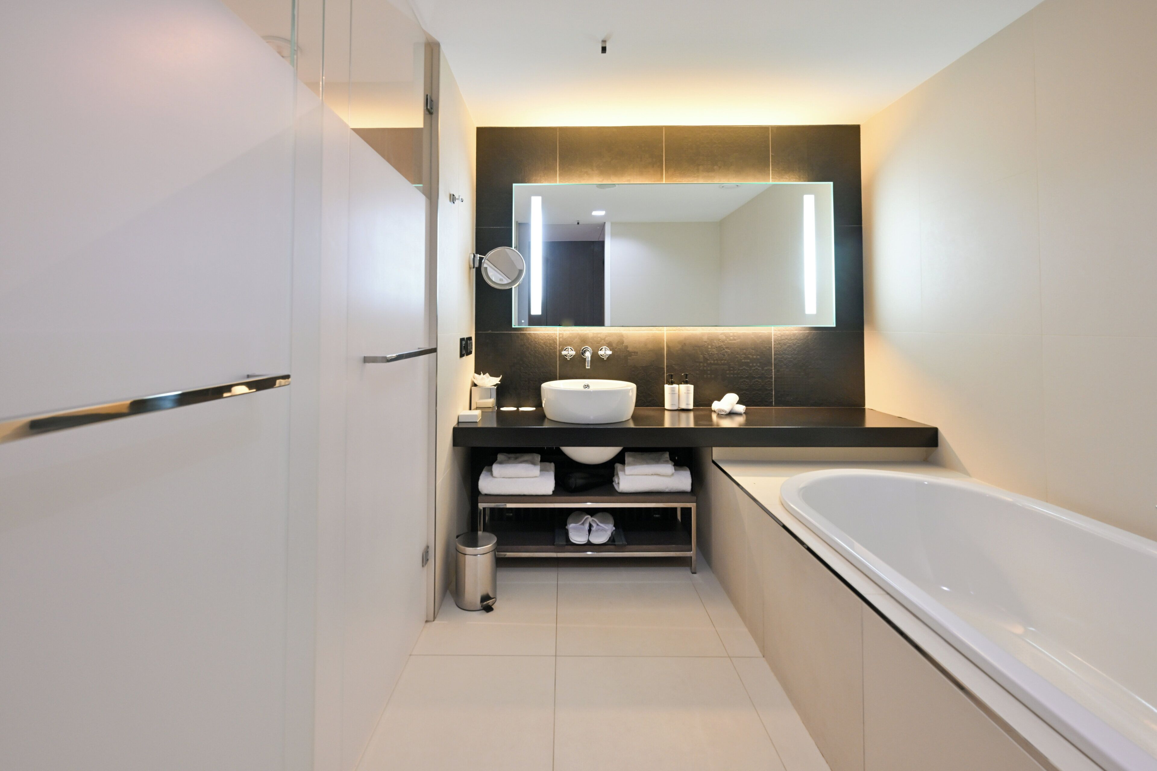 A modern bathroom with a large bathtub, a white sink, a mirror, and a shelf with towels.