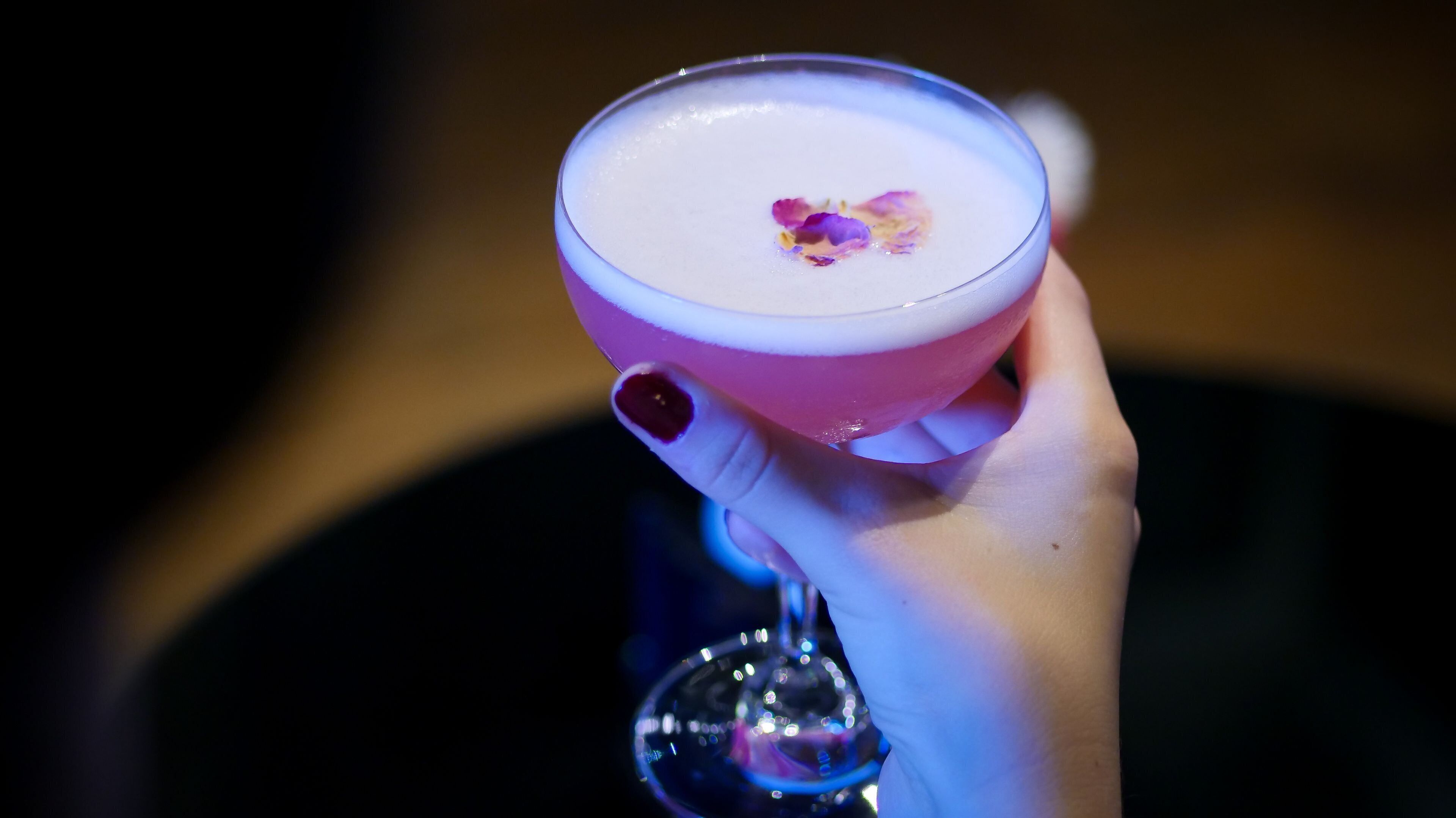 Eine Hand hält ein Cocktailglas mit rosafarbenem Rand und einer Blüten garnierung.