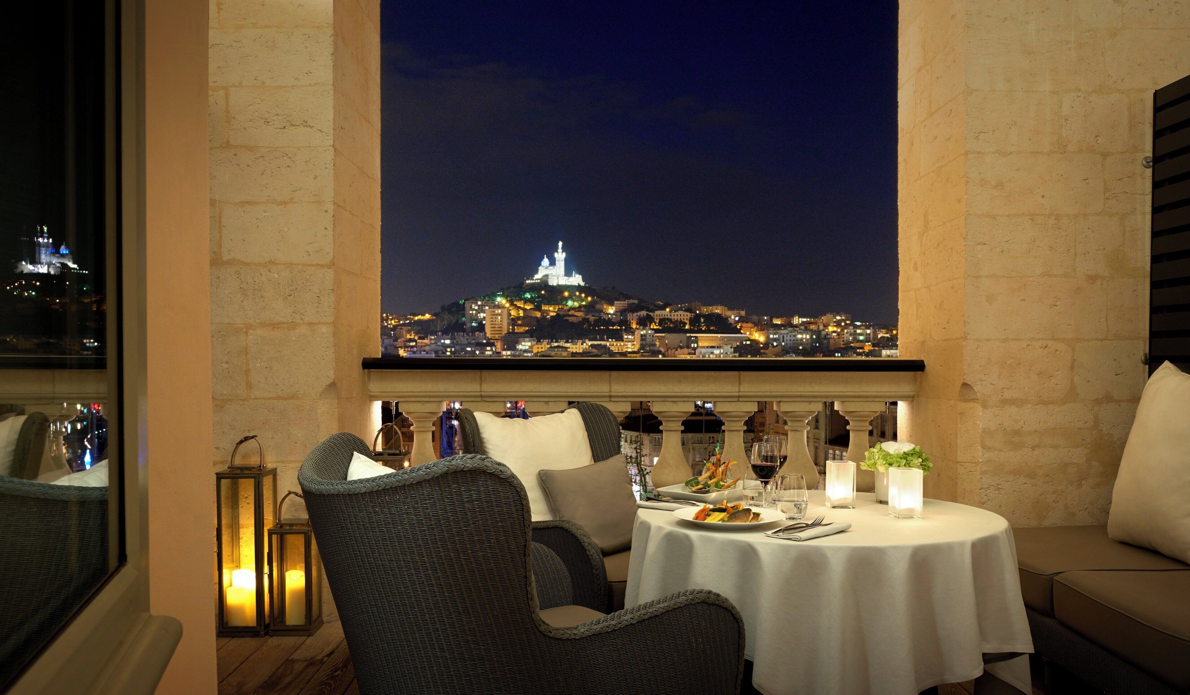 A dining area with wicker furniture, a table set for two, and a view of a cityscape at night.