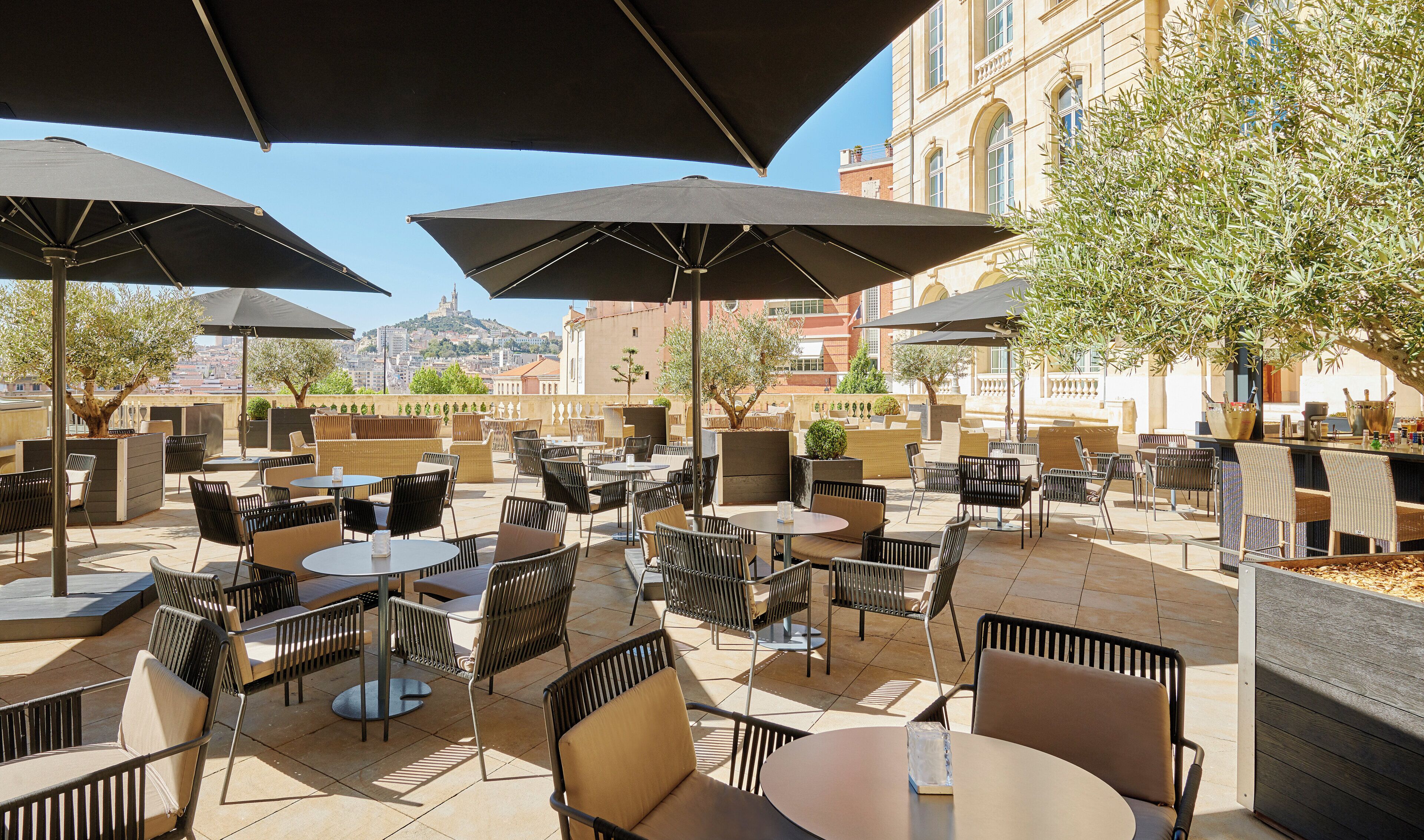 An outdoor café with tables and chairs, umbrellas, and a view of a cityscape.