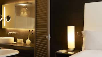 Quarto de hotel moderno com uma cama grande, mesa de cabeceira com abajur, banheiro com pia e espelho, e bancada com artigos de higiene pessoal.