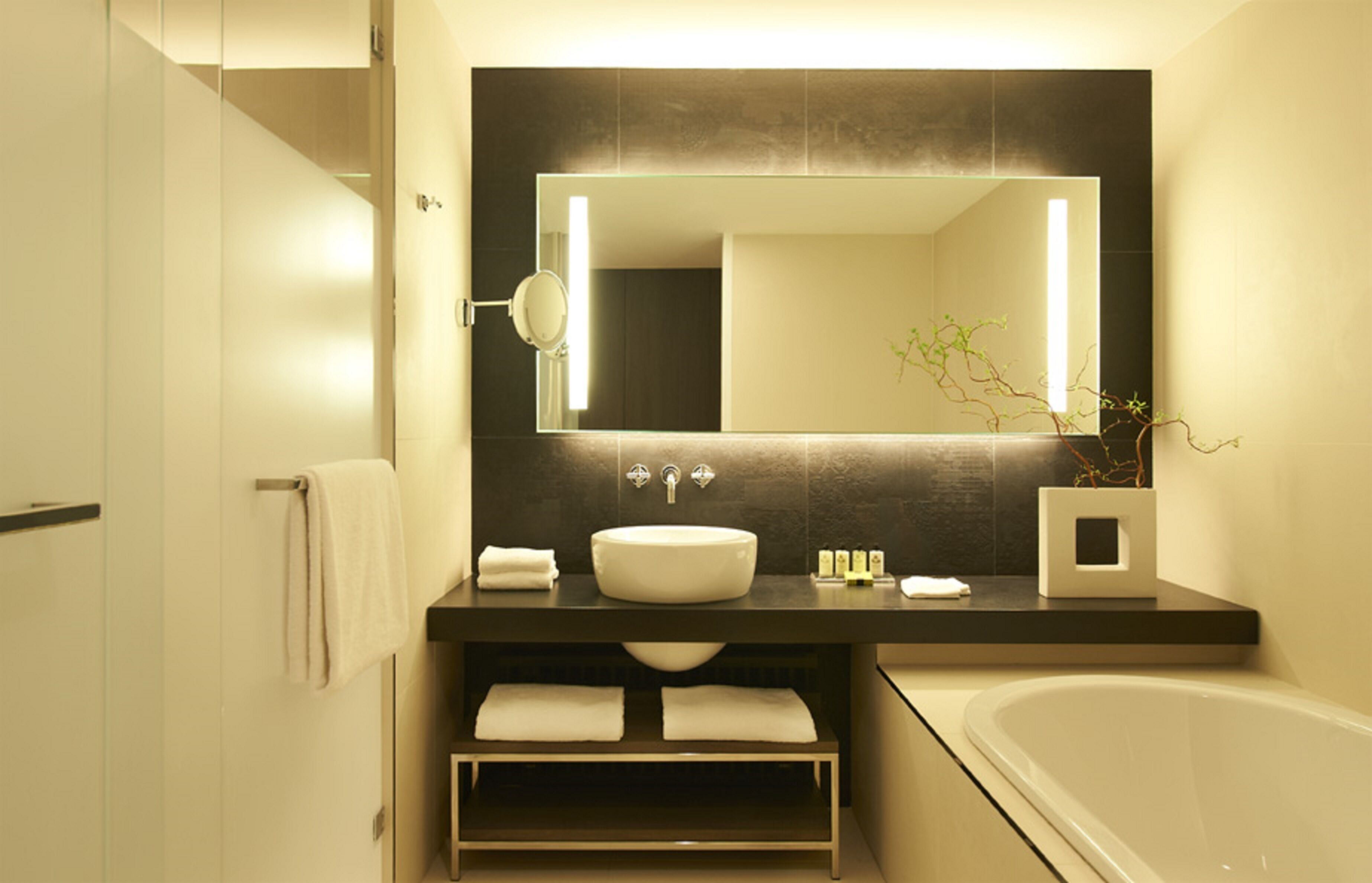 Ein modernes Badezimmer mit einem großen Spiegel, einem weißen Waschbecken, einer Badewanne und einem Regal mit Handtüchern.