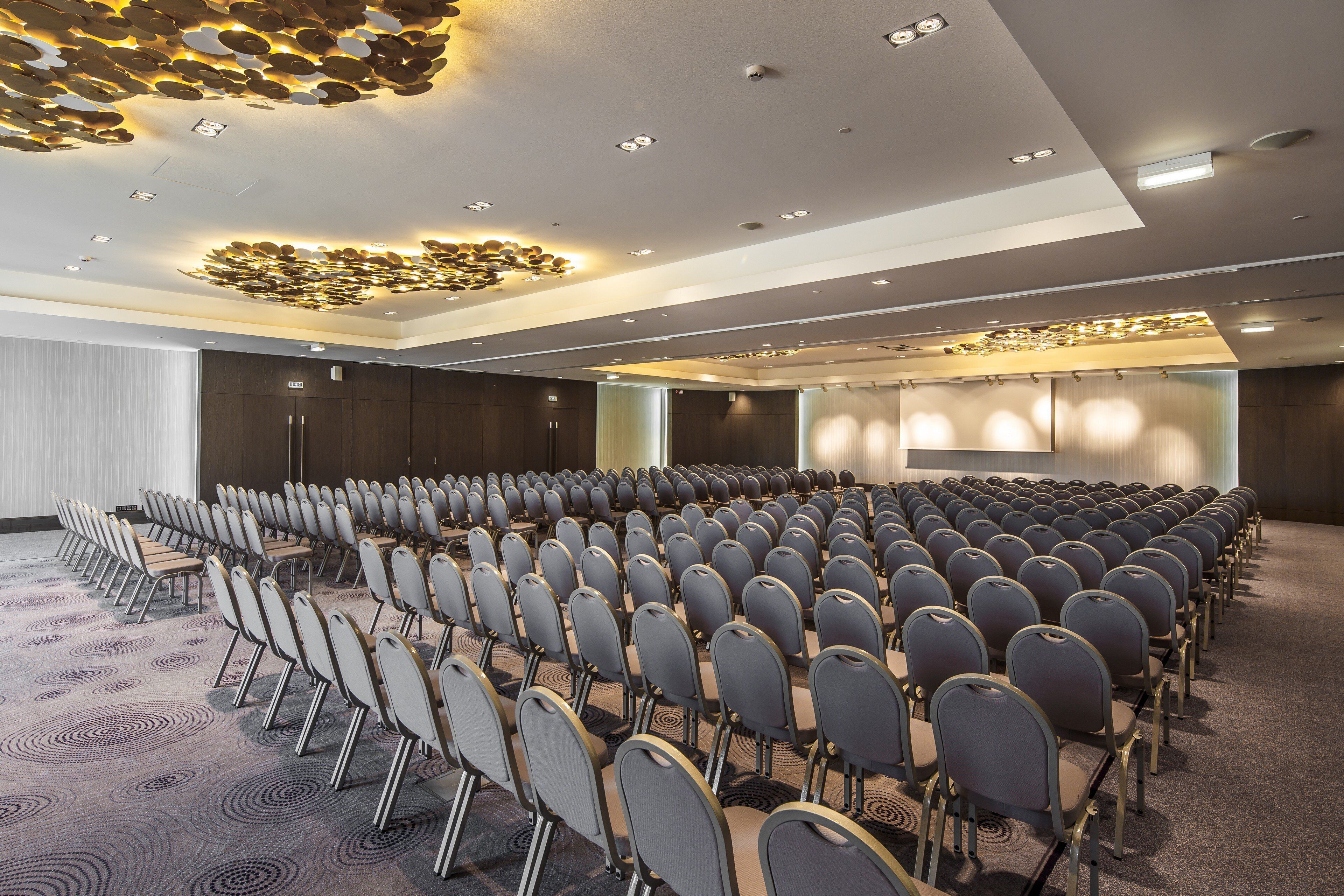 Une salle de conférence avec des rangées de chaises, une scène et un design moderne au niveau du plafond.