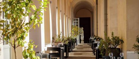 Un couloir avec des tables et des chaises, des plantes en pot et une vue sur une pièce.