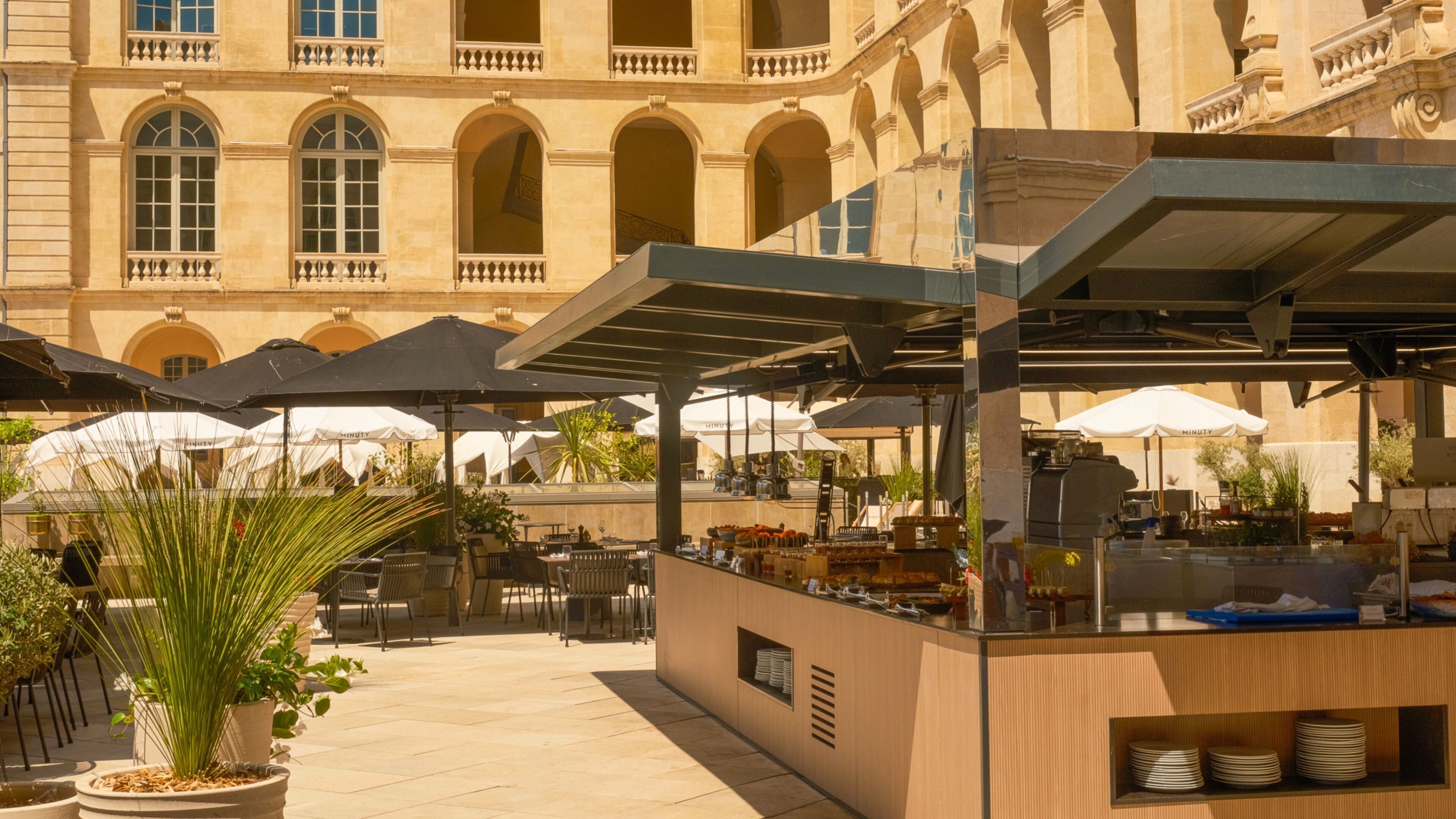 An outdoor café with tables and umbrellas, a food stall with a counter, and a building with arches in the background.