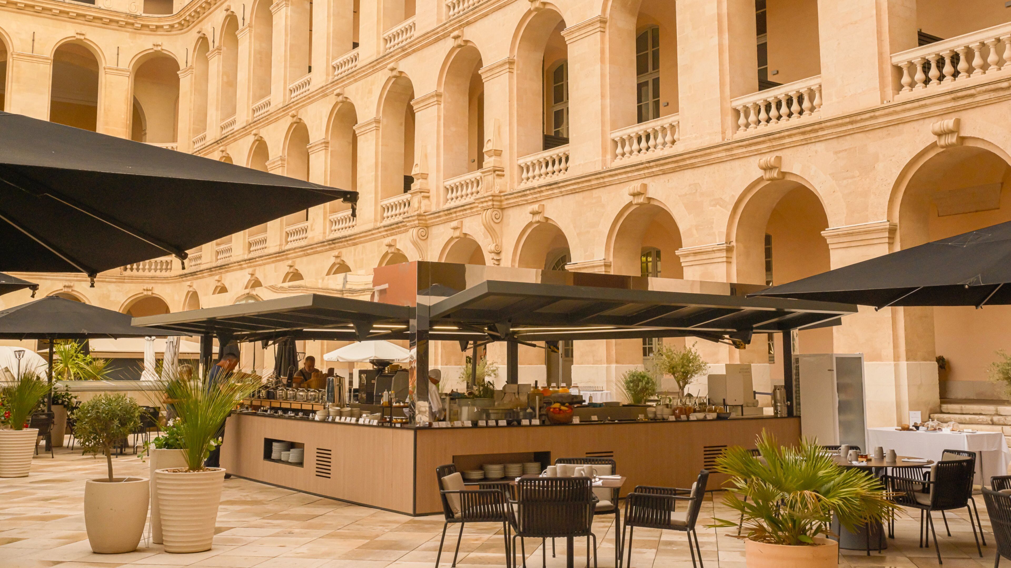 An outdoor café with tables and umbrellas, set against a building with arches and columns.