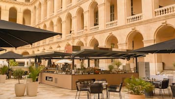 An outdoor café with tables and umbrellas, set against a building with arches and columns.