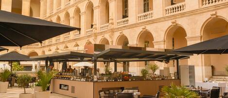 An outdoor café with tables and umbrellas, set against a building with arches and columns.