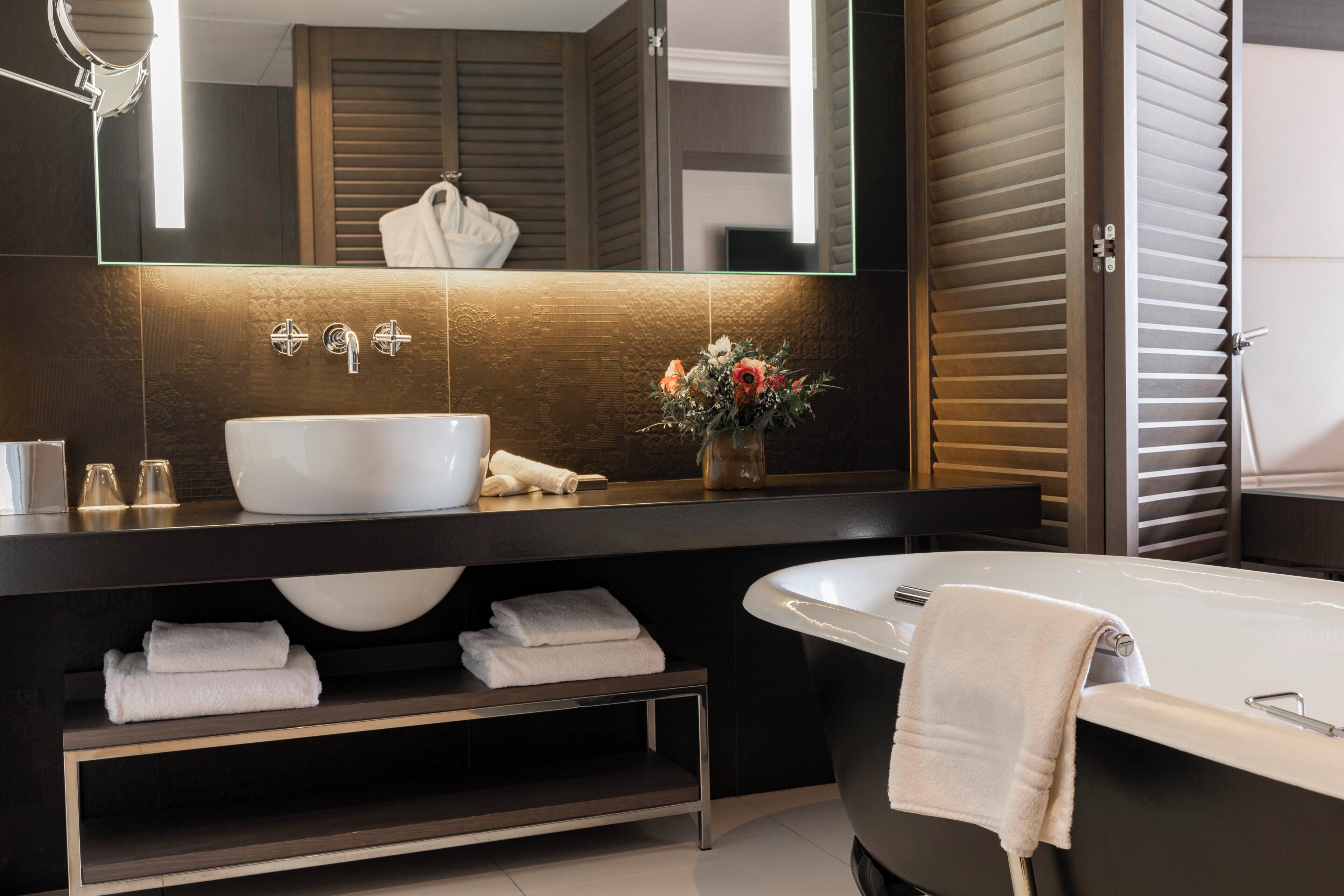 A modern bathroom with a large white sink, a bathtub, a mirror, and a shelf with folded towels.