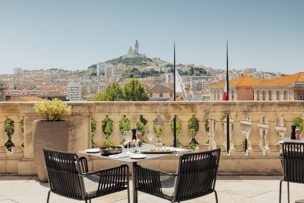 A rooftop terrace with a table set for dining, chairs, and a view of a cityscape with a hill and buildings.