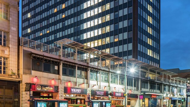 Un hôtel moderne, reconnaissable à son grand panneau « Hampton », qui s’étend sur plusieurs étages et dont le rez-de-chaussée abrite un espace commercial avec divers magasins.