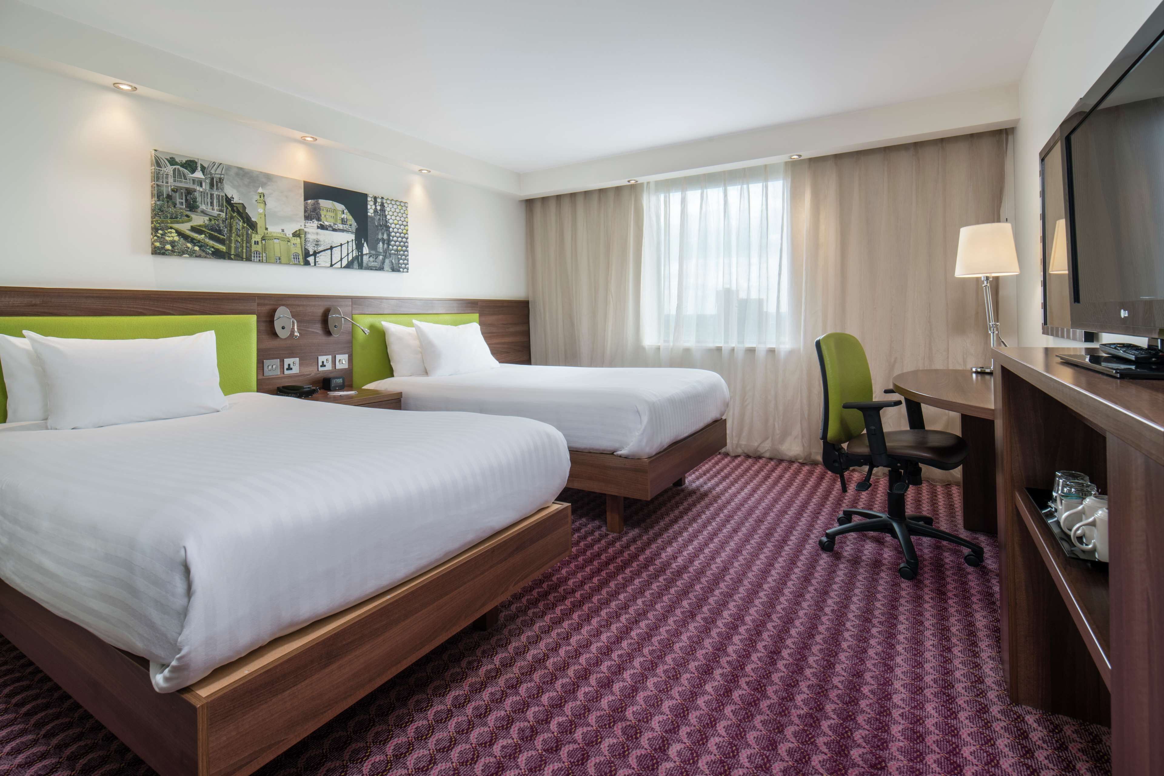 A hotel room with two beds, a desk, a chair, a TV, and a painting on the wall.