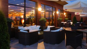 An outdoor seating area with wicker furniture and umbrellas, large windows, and indoor lighting visible through the glass. - Odyssey ClubHotel Wellness & SPA (Maslów)