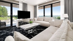 A modern living room with a large flat-screen TV, a sofa, a coffee table, and a view of a green landscape through large windows. - Odyssey ClubHotel Wellness & SPA (Maslów)
