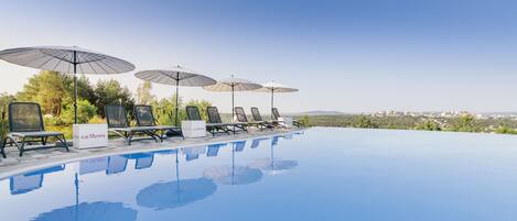Ein Infinity-Pool mit Liegestühlen und Sonnenschirmen, ein Schild für GH Mumm sowie ein Blick auf die Stadt im Hintergrund.